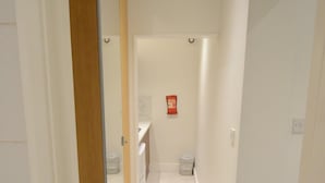 A narrow hallway with white walls, a wooden door frame, and a tiled floor. A small trash bin is visible in the distance. - Amber Residence Hotel (London)