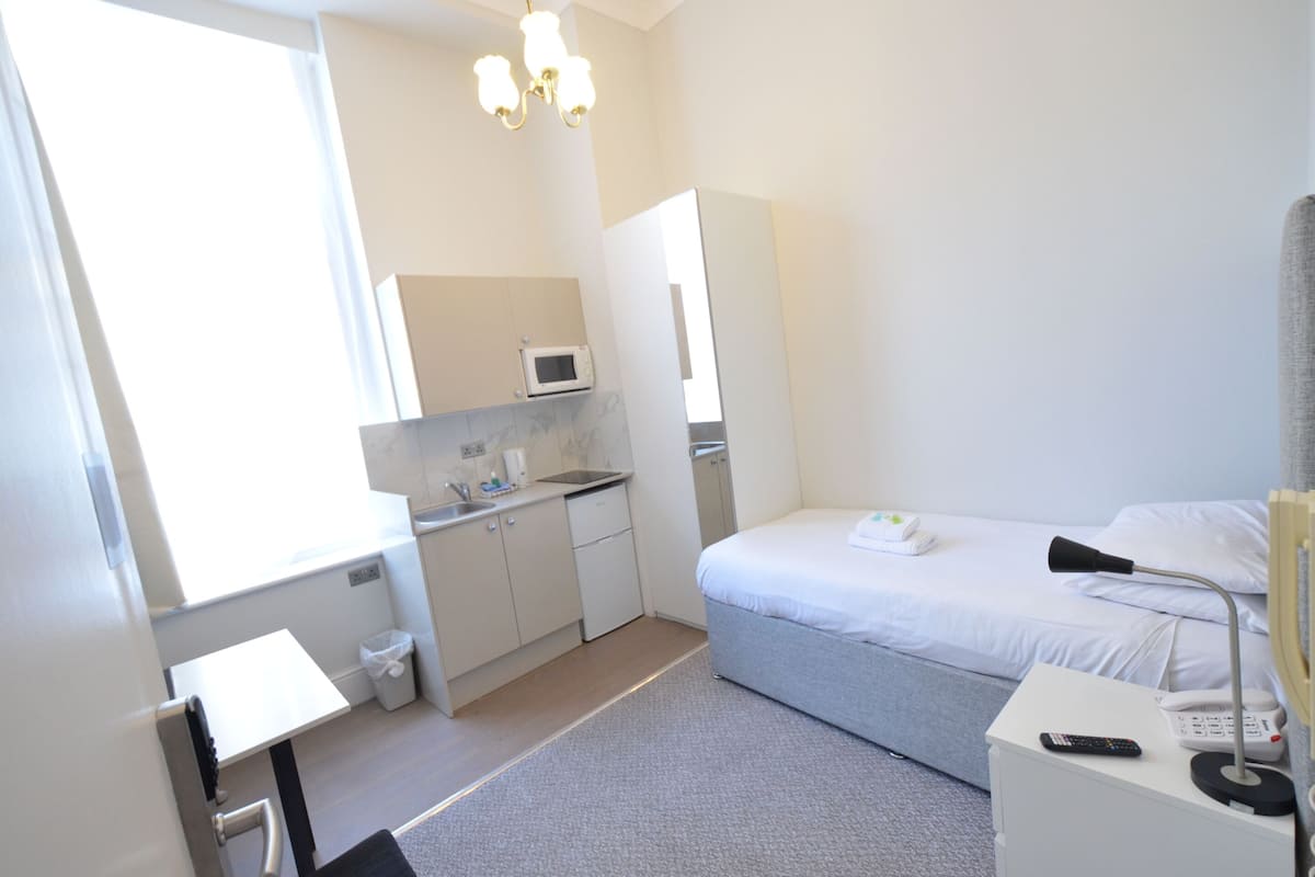 A compact hotel room with a kitchenette, a bed, a bedside table with a lamp, and a small table with a remote control.