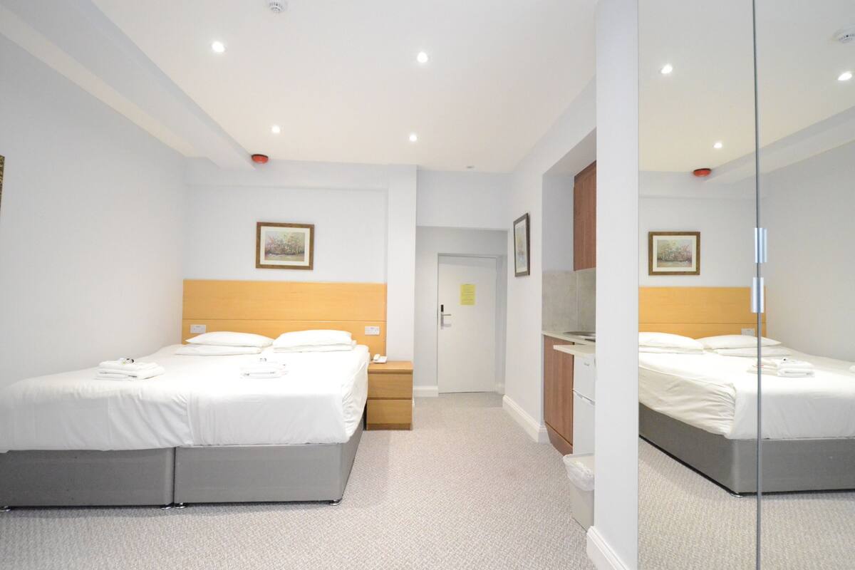 A hotel room with two beds, a wooden headboard, a bedside table, a mirror, and a door leading to a bathroom.