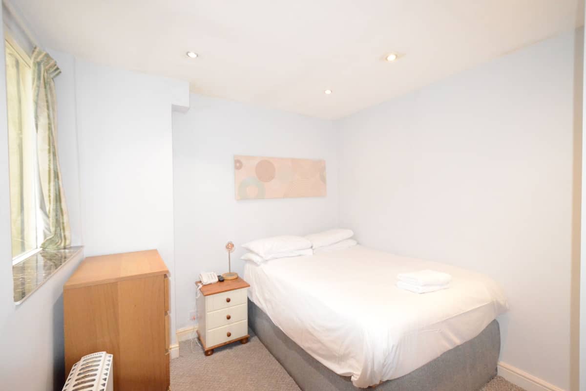 A bedroom with a single bed, a bedside table with a lamp, a wooden cabinet, and a window with curtains.
