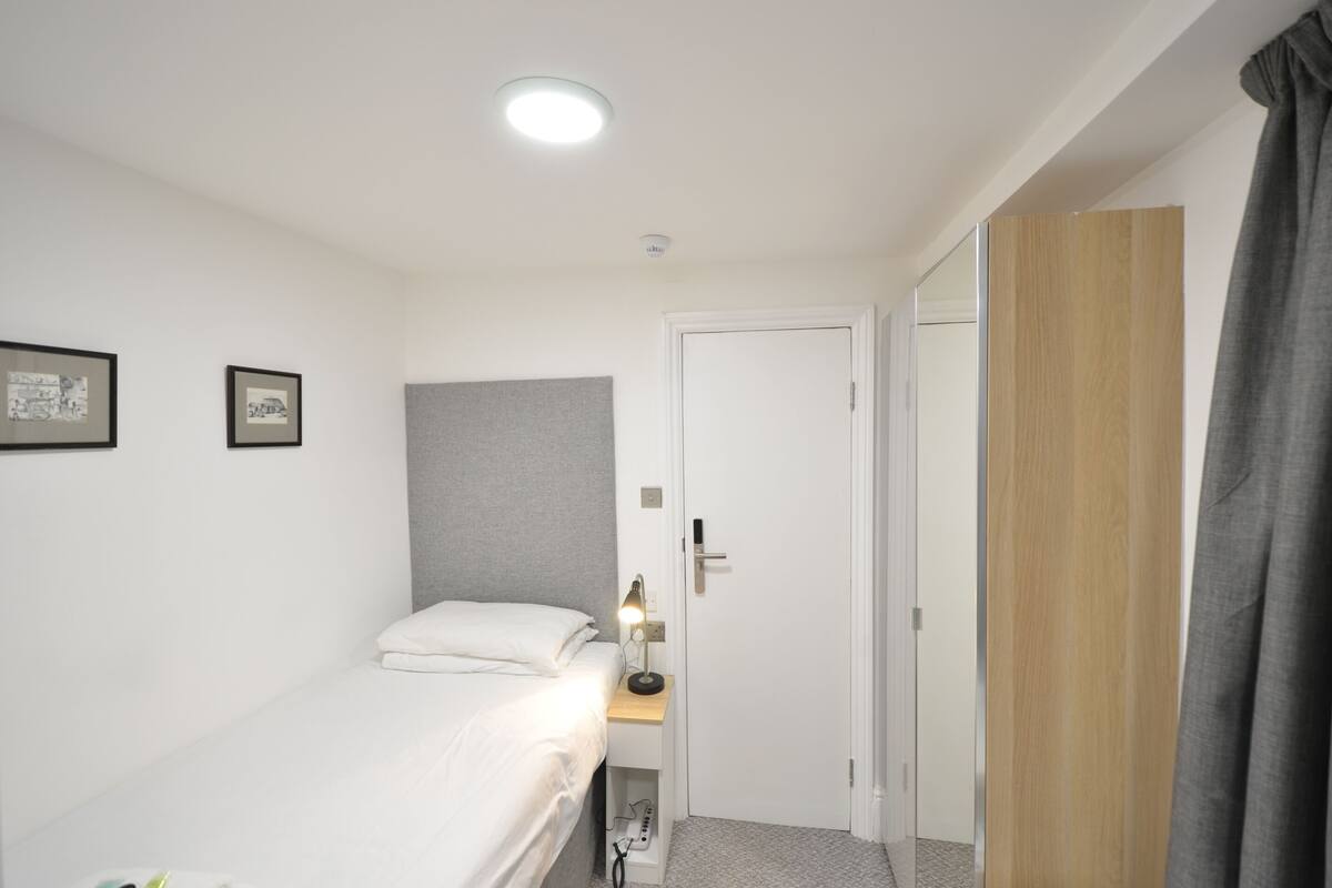 A small, neatly organized bedroom with a single bed, a bedside table with a lamp, a door, and a wardrobe.