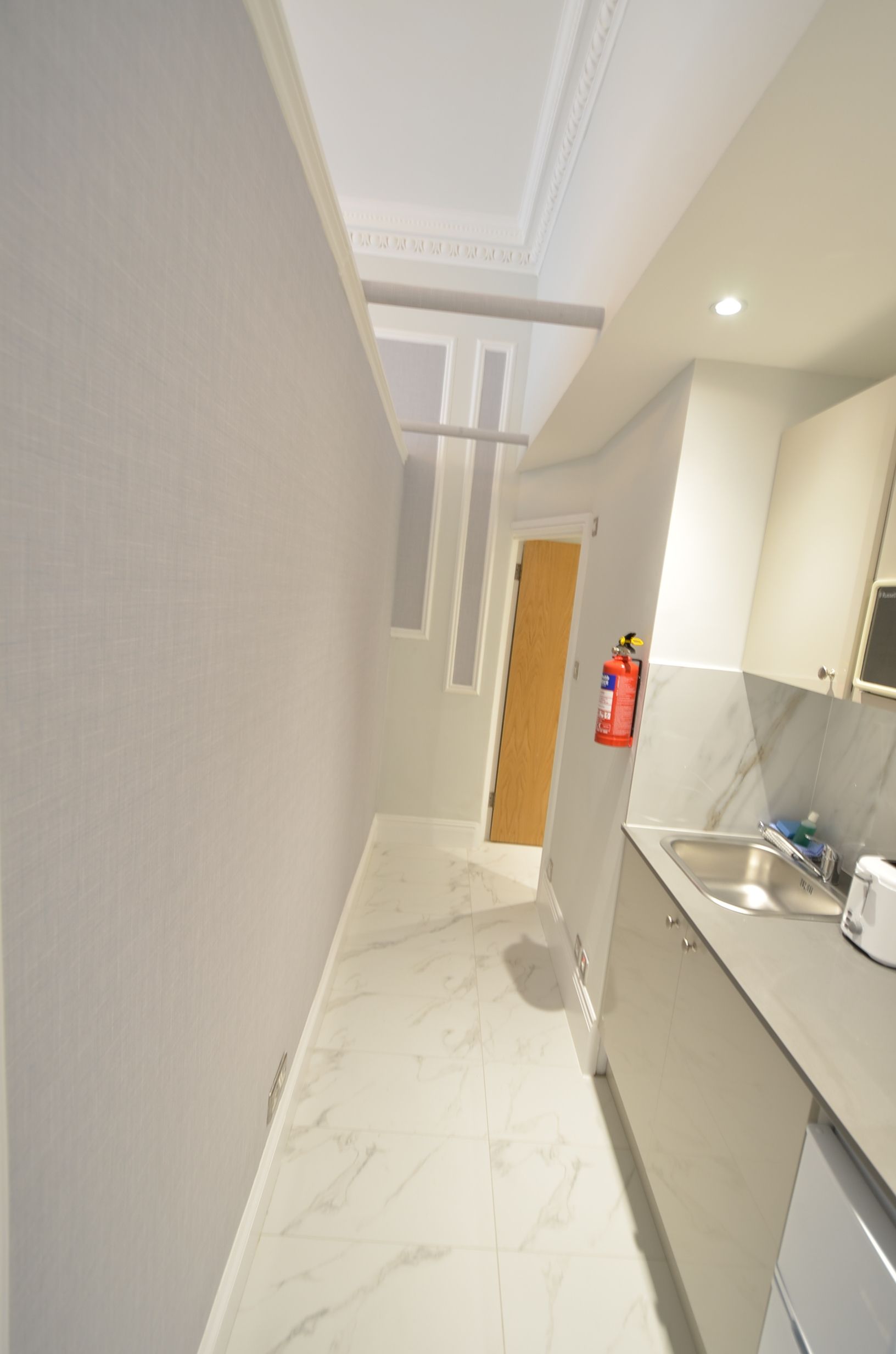 A narrow hallway with a kitchen area on the right. The hallway has a white tiled floor and walls, and a wooden door at the end.