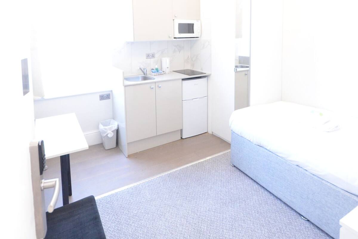 A small, bright room with a kitchenette, a bed, and a desk.