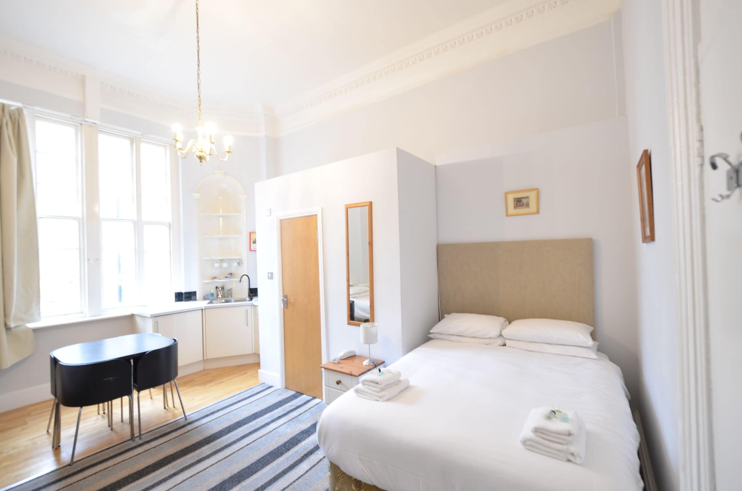 A hotel room with a bed, bedside table, and a small desk with a chair. There is a window with curtains, a chandelier, and a mirror on the wall.