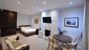 A modern hotel room with a bed, a desk, a chair, a TV, a fireplace, and a dining area. - Amber Residence Hotel (London)