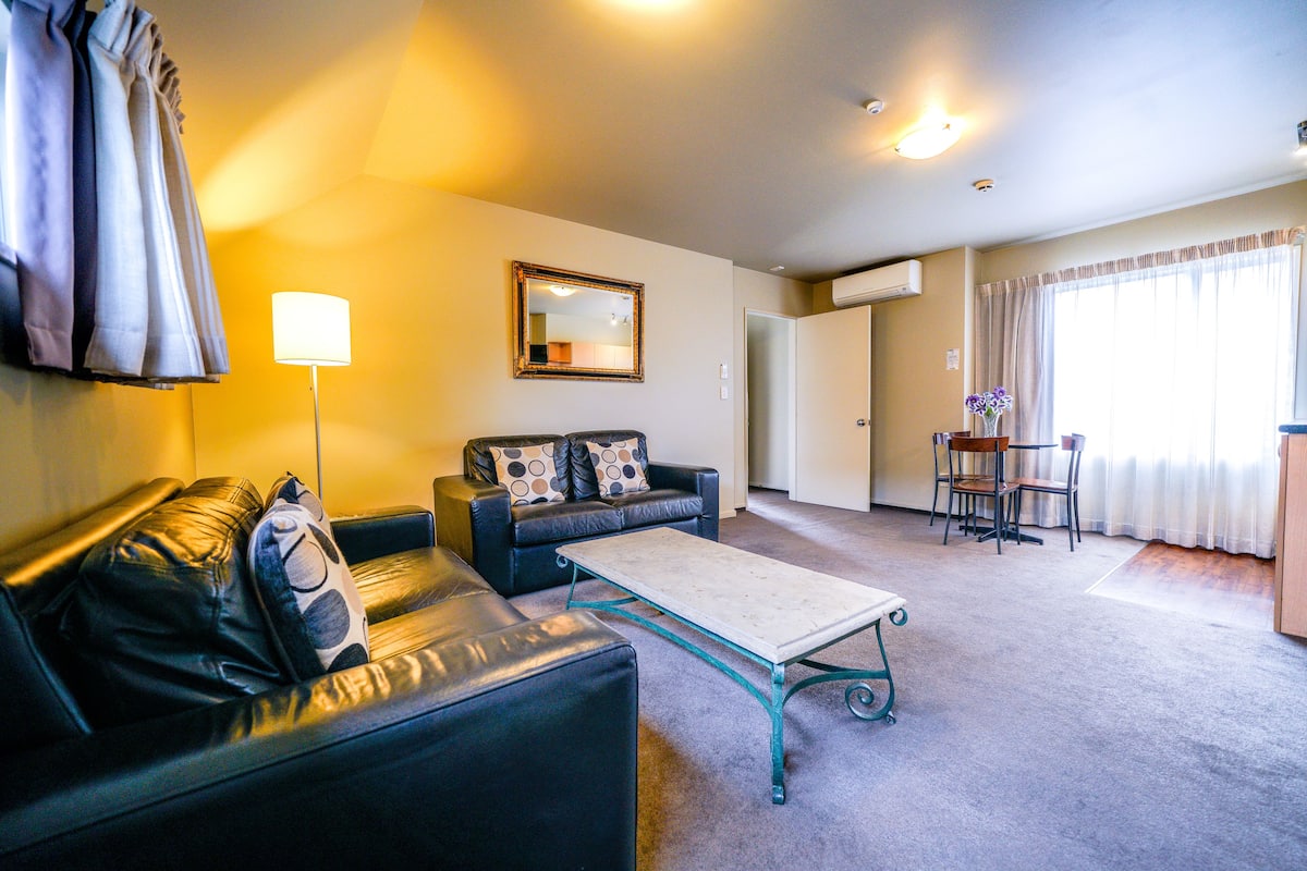 A living room with a sofa, a coffee table, a lamp, a mirror, and a dining area with chairs and a table.