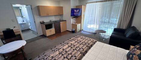 A hotel room with a bed, a desk, a chair, a TV, a kitchen area, and a bathroom.
