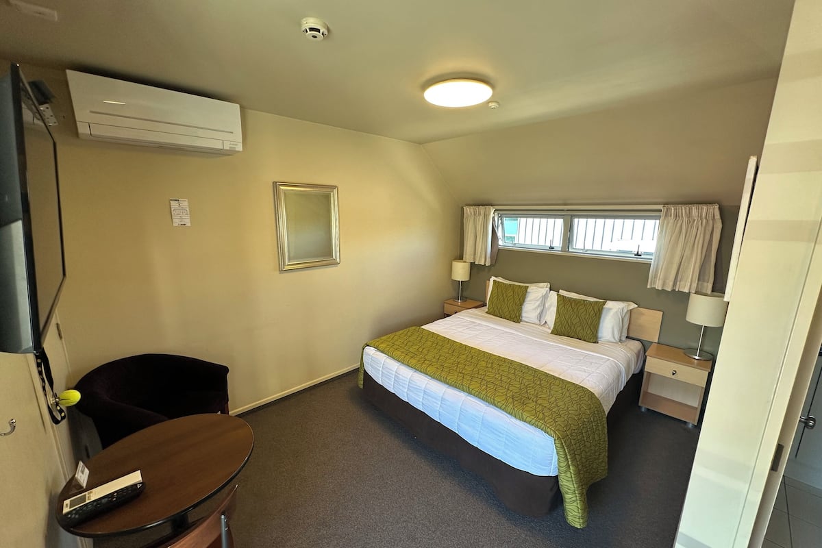 A hotel room with a bed, bedside tables, a chair, a small table, an air conditioning unit, a mirror, and a window with curtains.