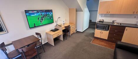 Una habitación de hotel pequeña con escritorio, silla, kitchenette y un televisor mostrando un partido deportivo.