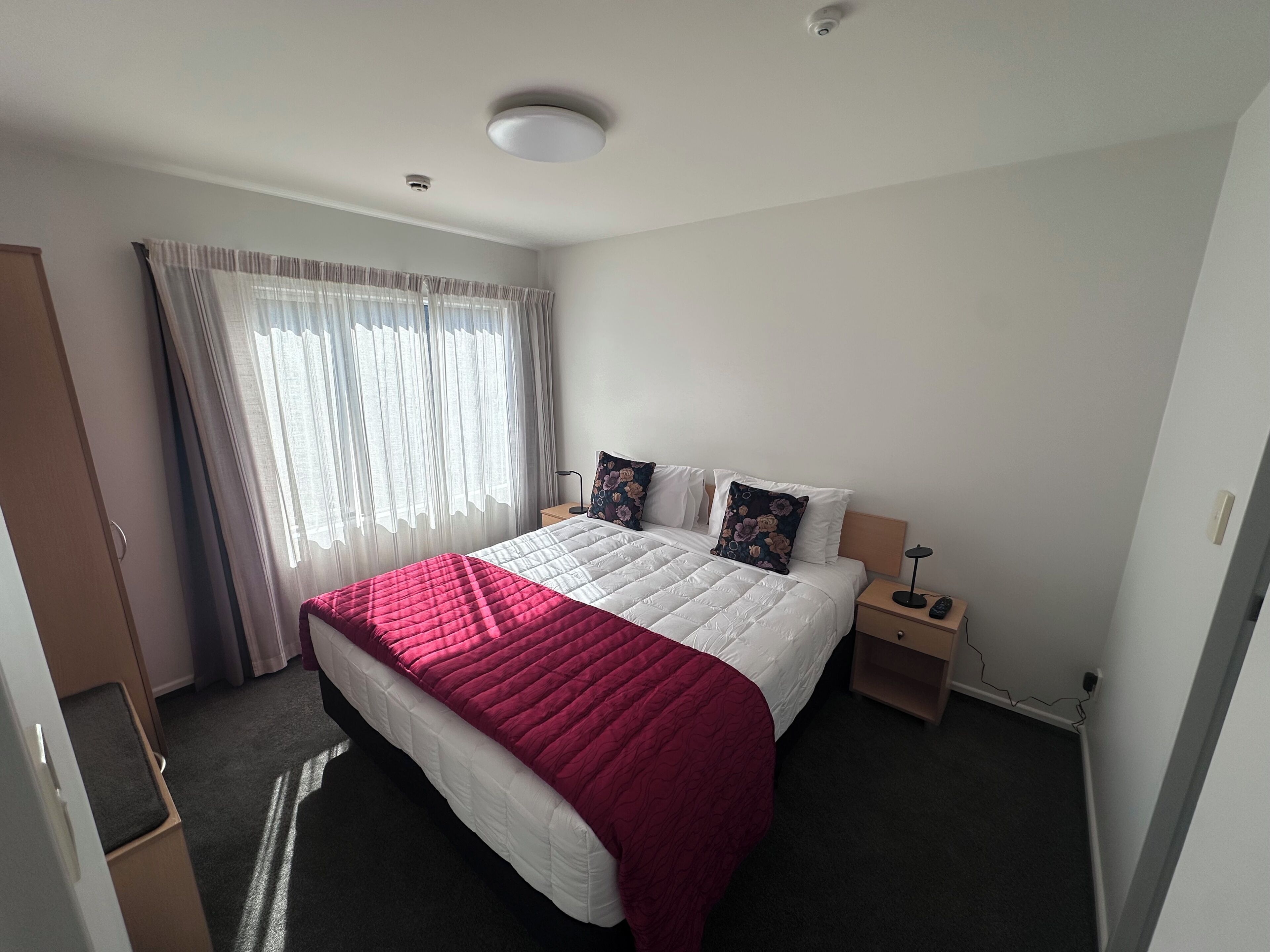 A hotel room with a bed, a nightstand, a lamp, a window with curtains, and a door.