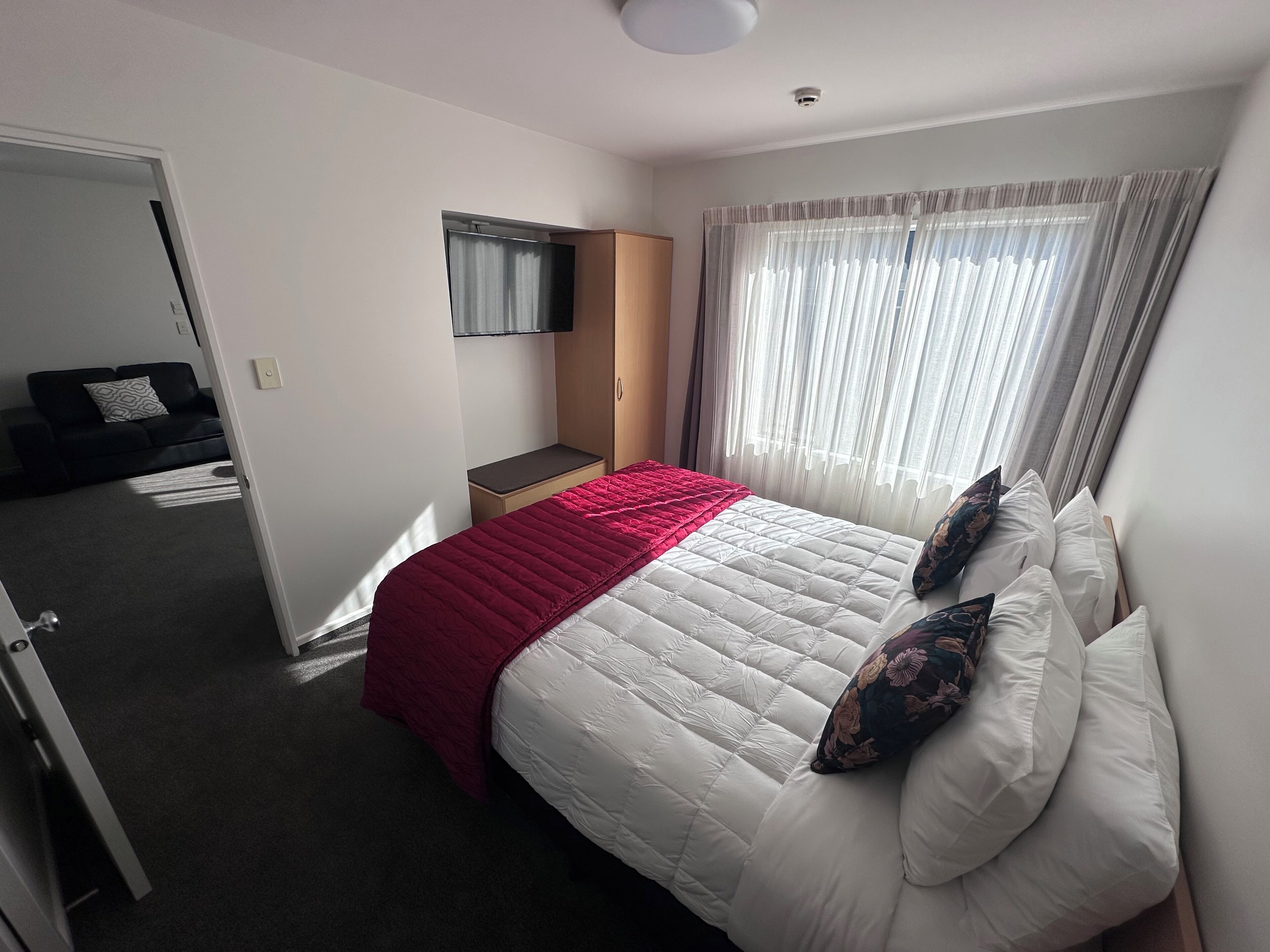 Una habitación de hotel con cama, televisor montado en la pared, ventana con cortinas y puerta que da a otra habitación.