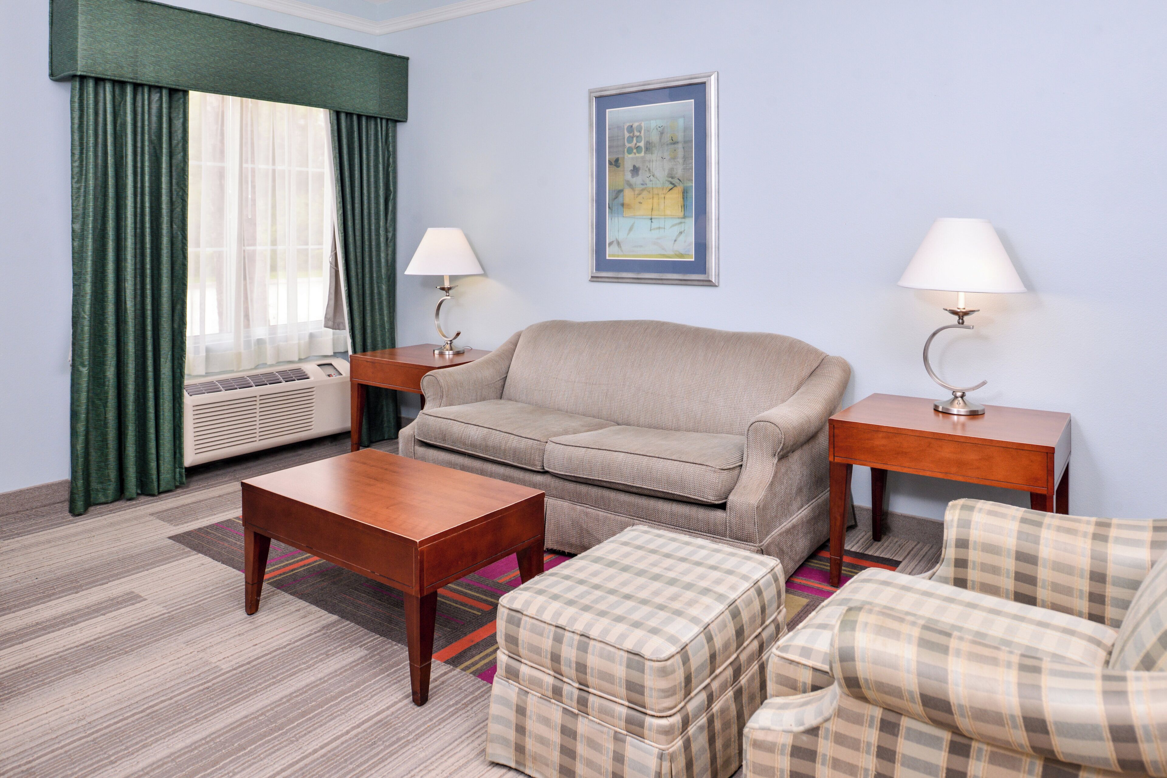 A hotel room with a sofa, a table, a chair, a small ottoman, a standing lamp, a bedside table, a framed picture on the wall, and a window with curtains.