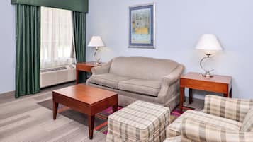 A hotel room with a sofa, a table, a chair, a small ottoman, a standing lamp, a bedside table, a framed picture on the wall, and a window with curtains.