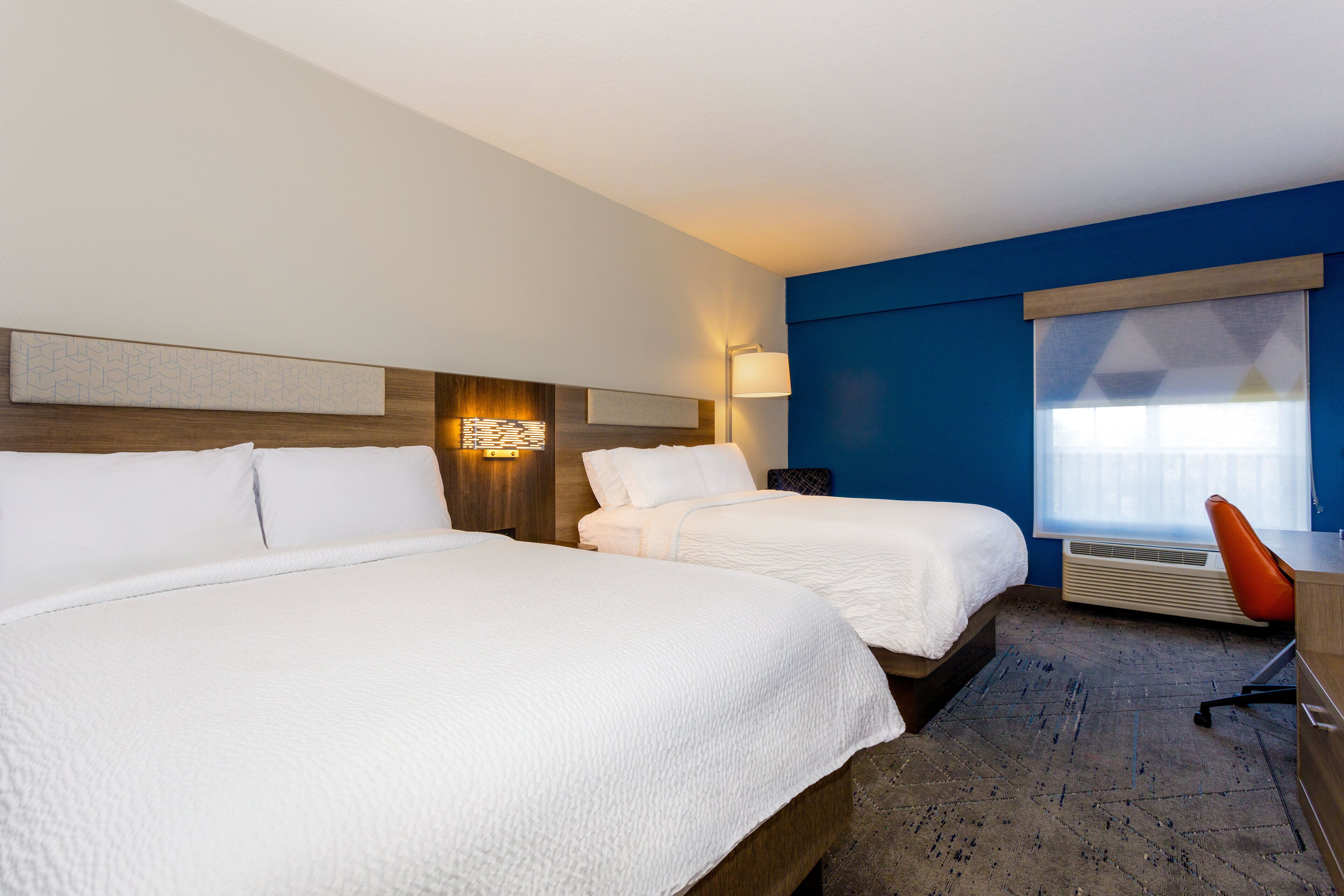 A hotel room with two beds, a desk, a chair, a lamp, and a large window.