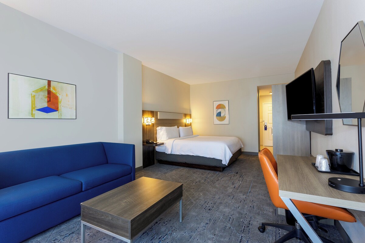 Hotelkamer met een blauwe bank, een houten bijzettafeltje, een oranje bureaustoel, een bed met witte lakens, een flatscreen-tv en kunst aan de muren.