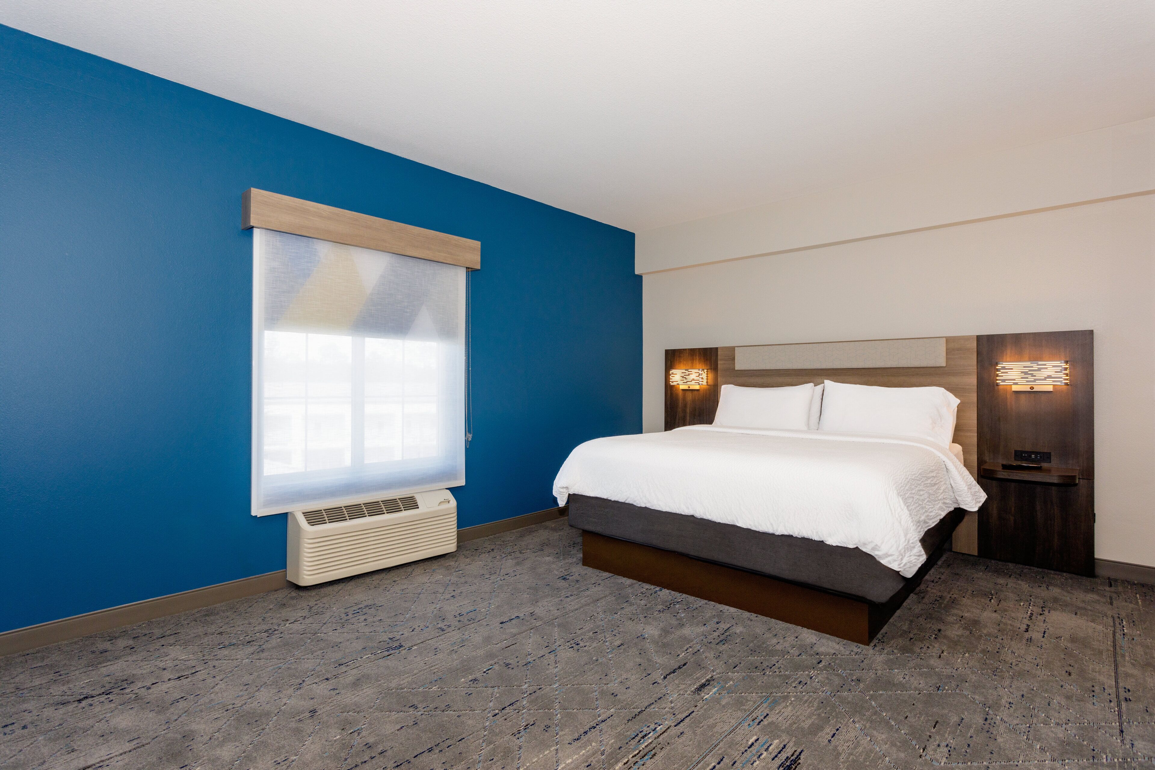 A modern hotel room with a large bed, a window with blinds, a wall-mounted heater, and a nightstand with a lamp.
