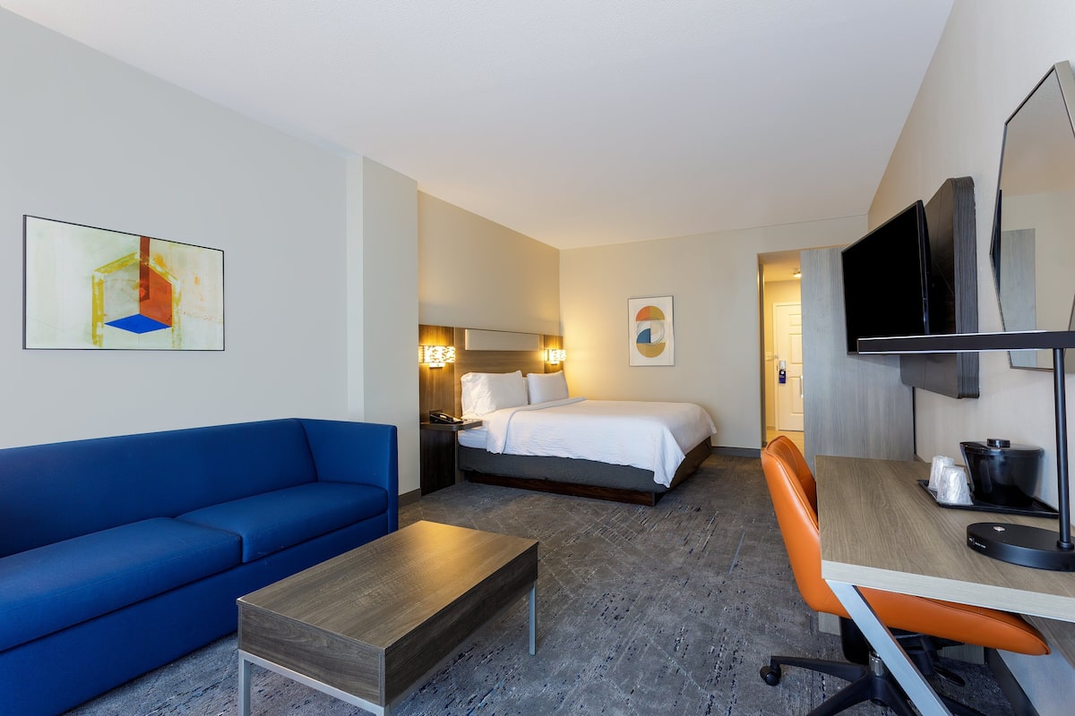 Habitación de hotel con un sofá azul, una mesa de centro de madera, una silla de oficina naranja, una cama con ropa blanca, un televisor de pantalla plana y obras de arte en las paredes.