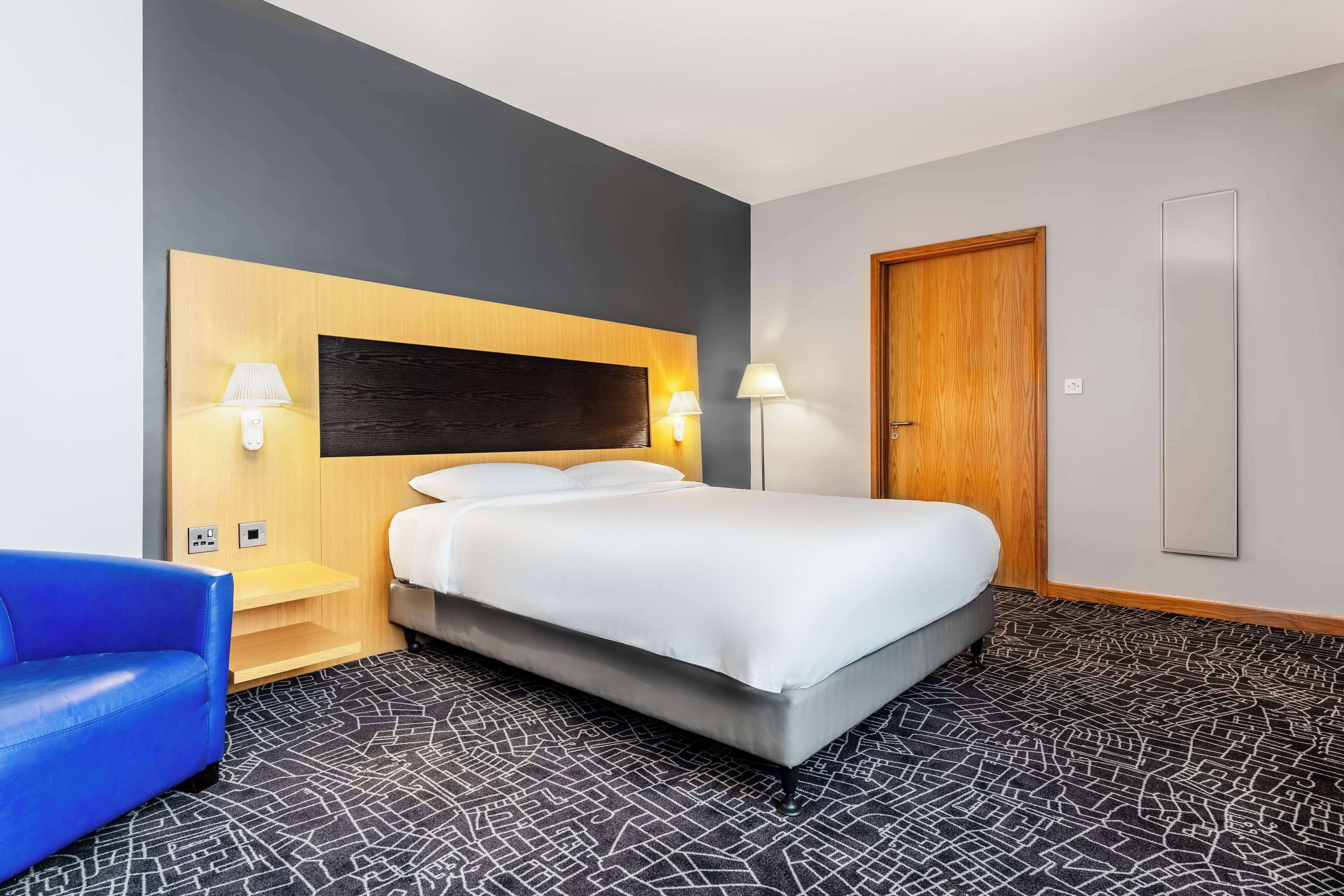 A hotel room with a bed, two bedside lamps, a blue chair, a wooden headboard, a door, and a patterned carpet.