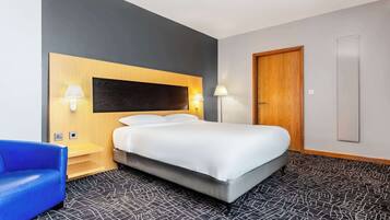 A hotel room with a bed, two bedside lamps, a blue chair, a wooden headboard, a door, and a patterned carpet.