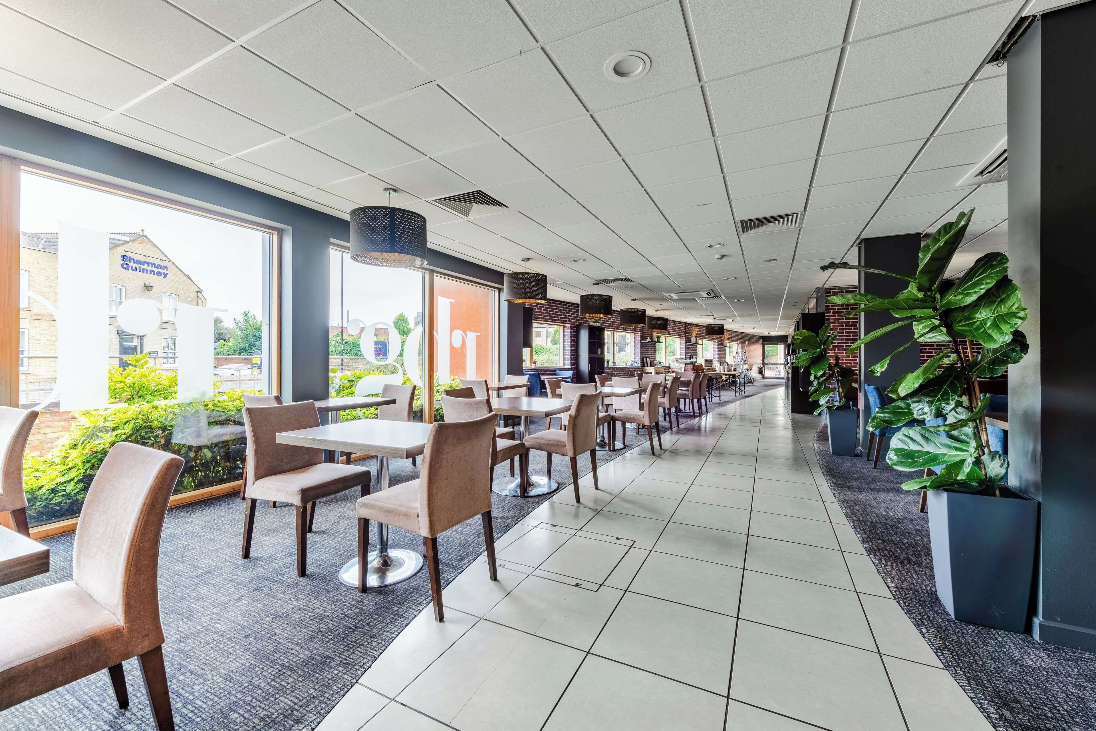 A spacious indoor area with multiple tables and chairs arranged in rows, large windows on one side, and potted plants along the walls.