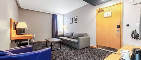 Habitación de hotel con un sofá gris, un sillón azul, una mesita, una lámpara, una puerta de madera y una zona de café.