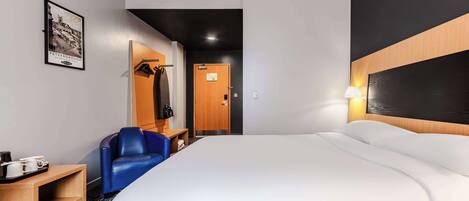 A hotel room with a large bed, a blue chair, a wooden side table with a tray of items, and a framed picture on the wall.