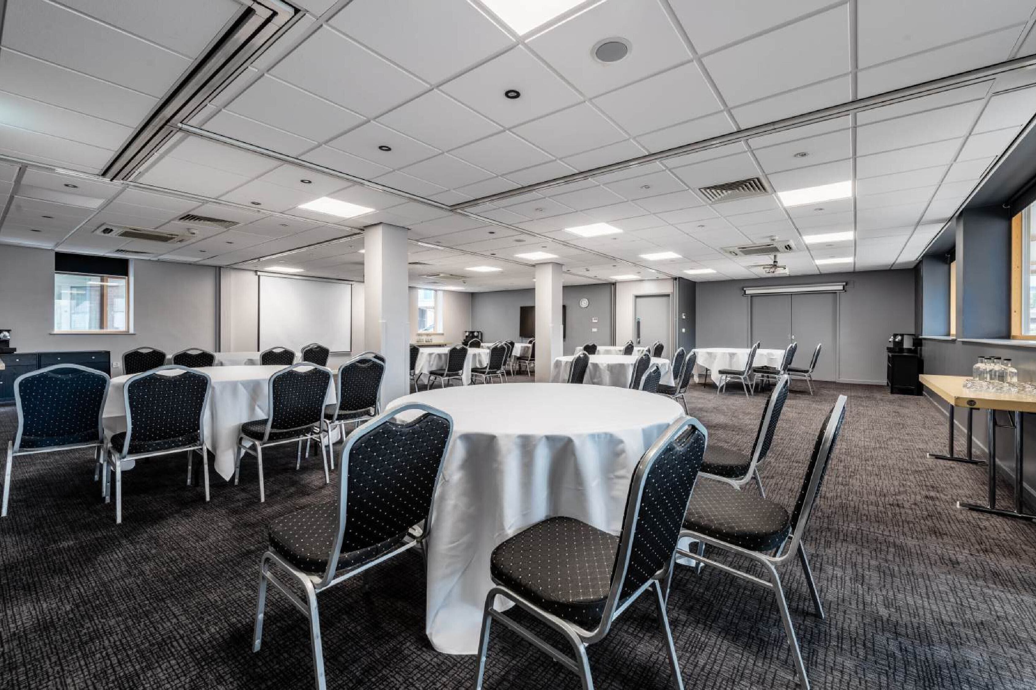 A conference room with round tables and chairs, carpeted floors, and a white ceiling with lighting fixtures.