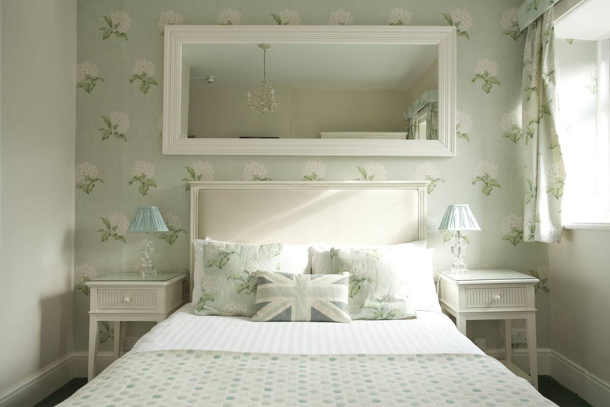 A neatly arranged bedroom with a large bed, two bedside tables with lamps, a mirror above the bed, and floral wallpaper.