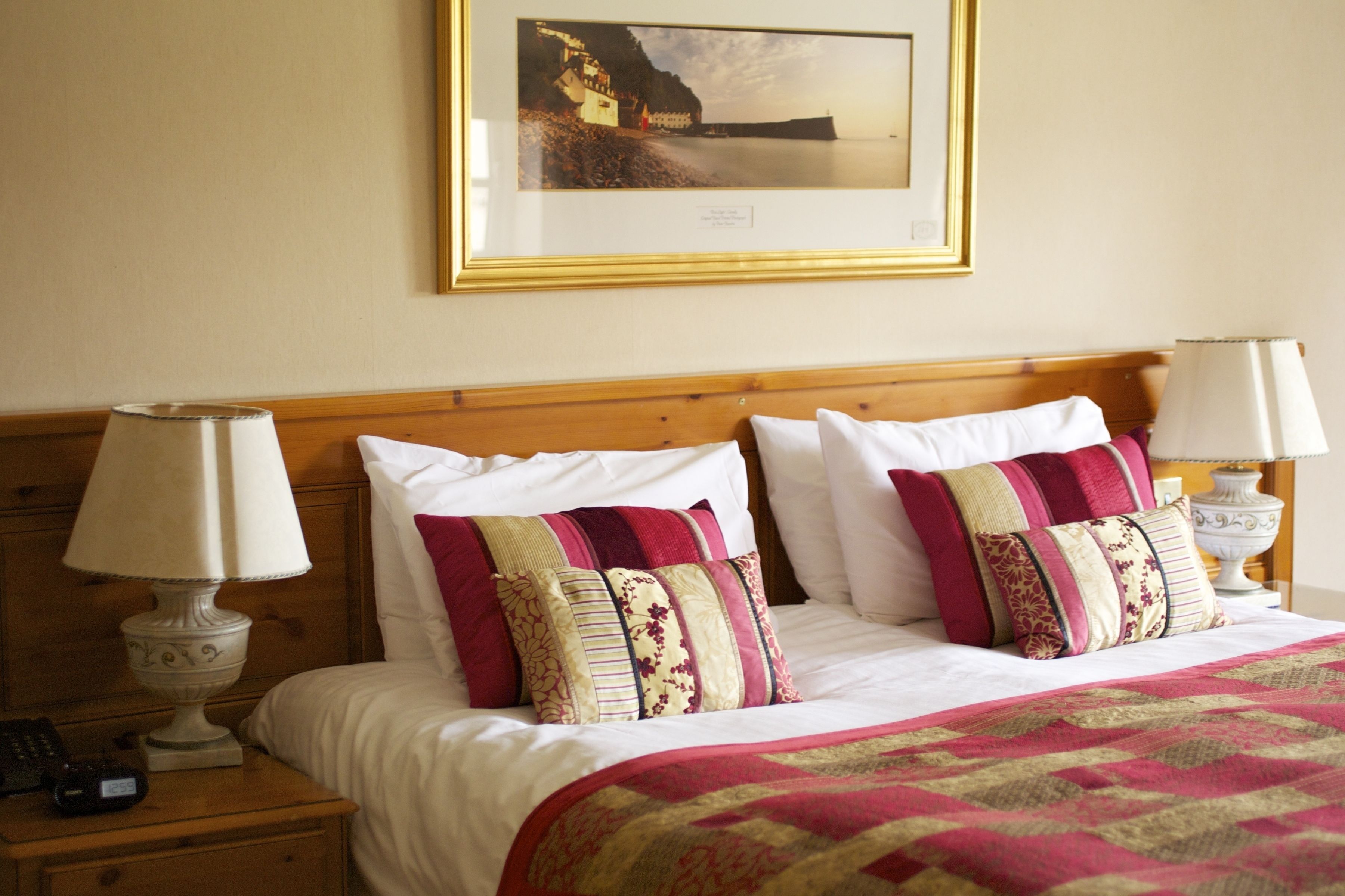Habitación de hotel con cabecera de madera, dos lámparas de noche, cama con ropa blanca y almohadas estampadas, un cuadro en la pared y un teléfono en la mesita de noche.