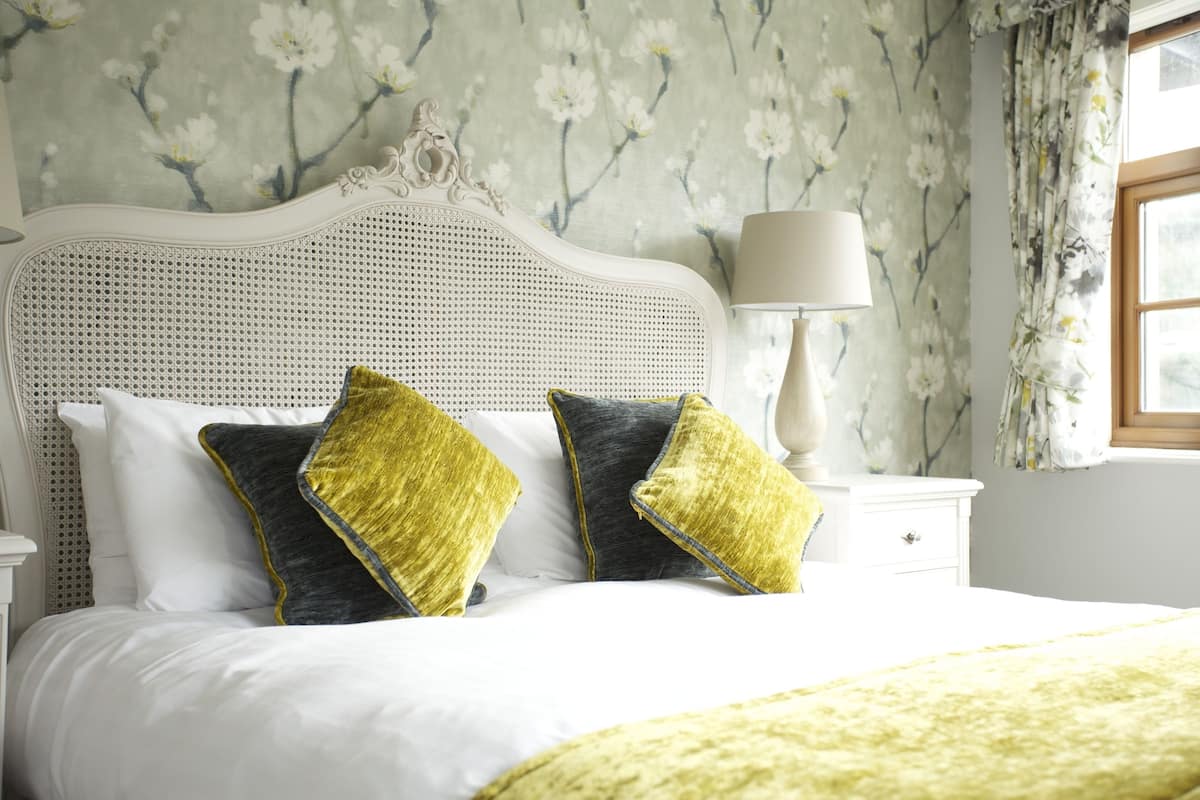 A neatly made bed with white bedding and two decorative pillows, a white bedside table with a lamp, and floral wallpaper.