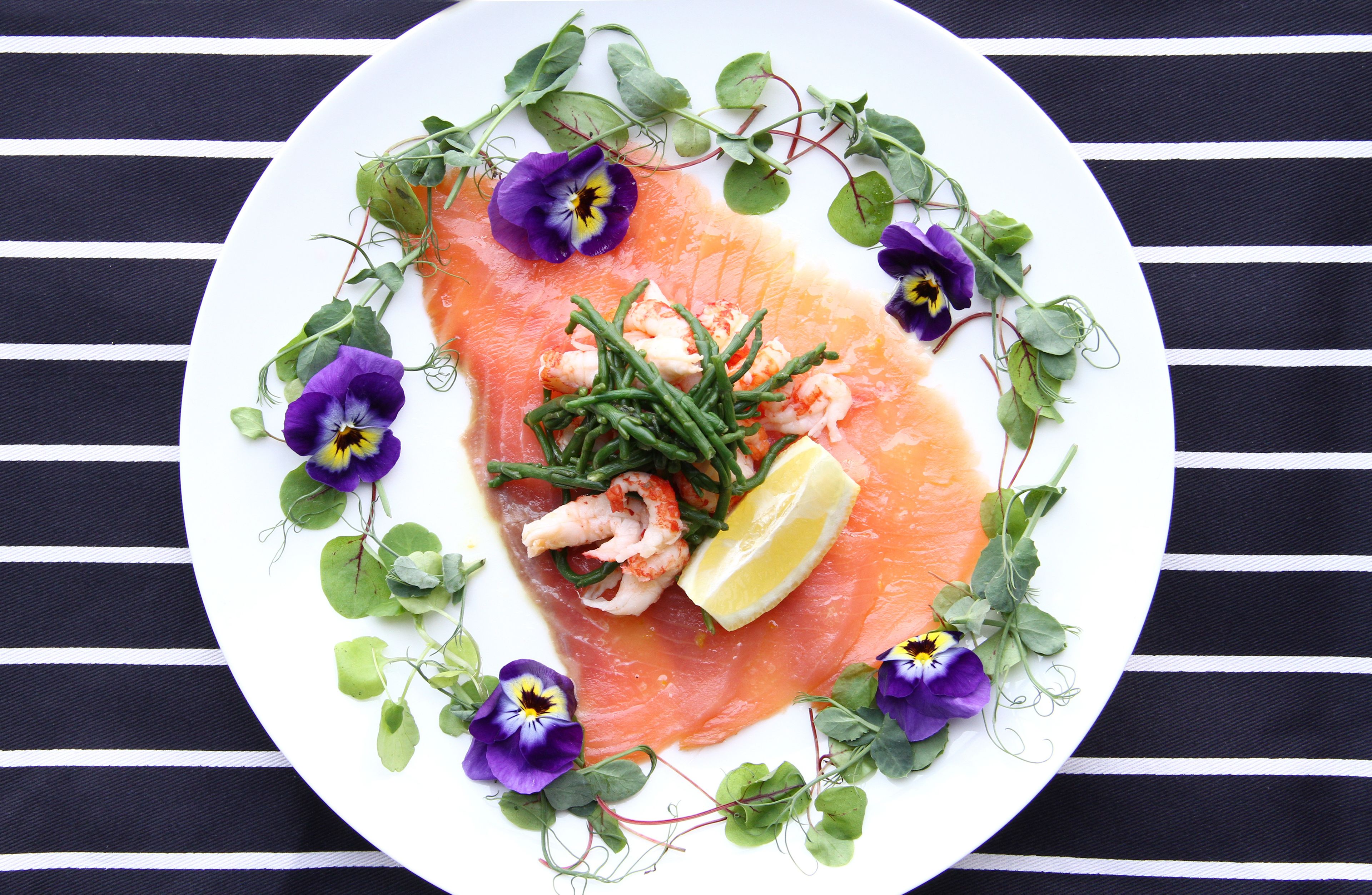 A plate of salmon with shrimp and greens, surrounded by purple flowers, on a white plate.