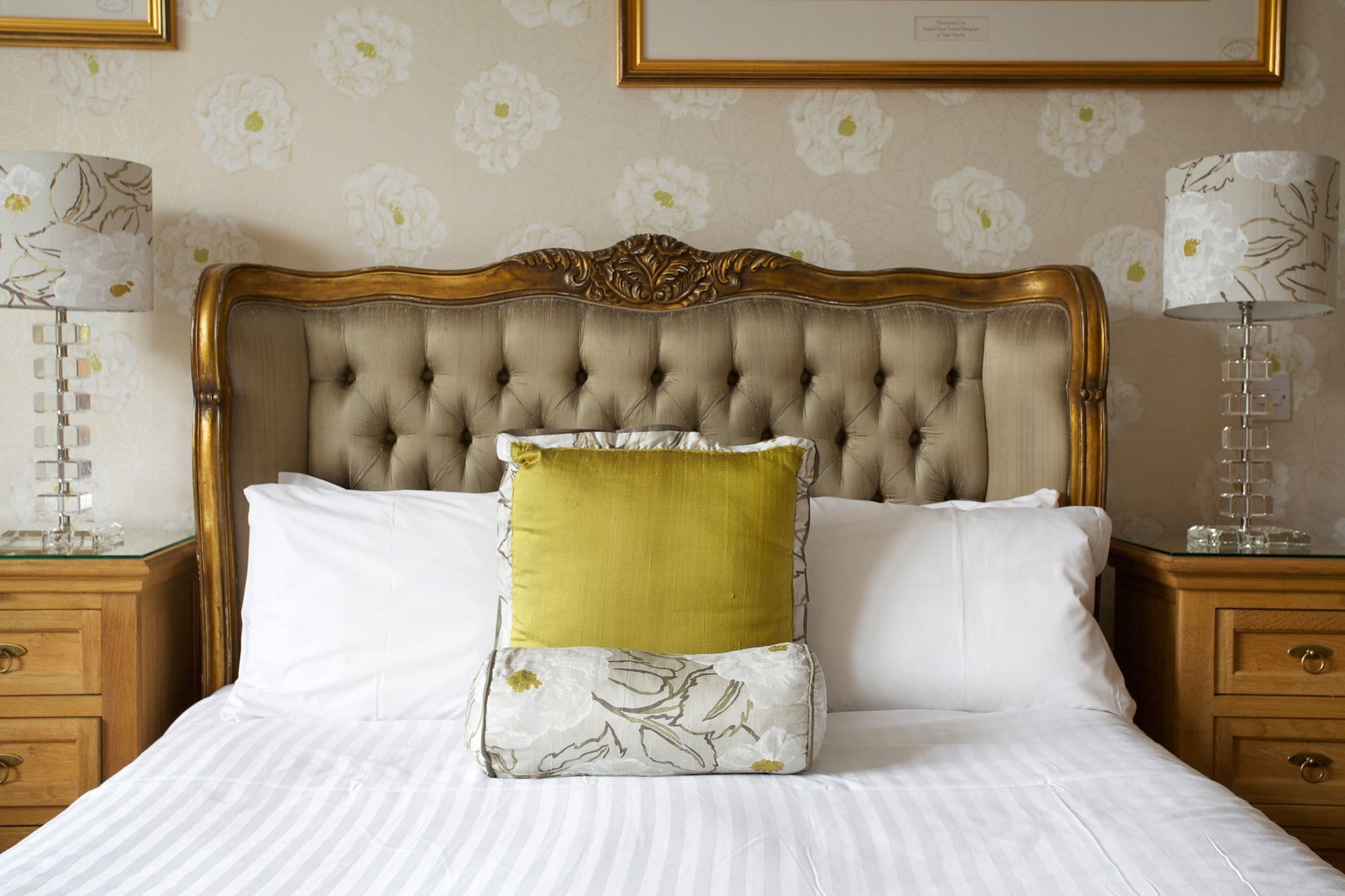 Un dormitorio con cabecera capitonada, una cama con almohadas blancas y amarillas, y mesitas de noche de madera con lámparas.