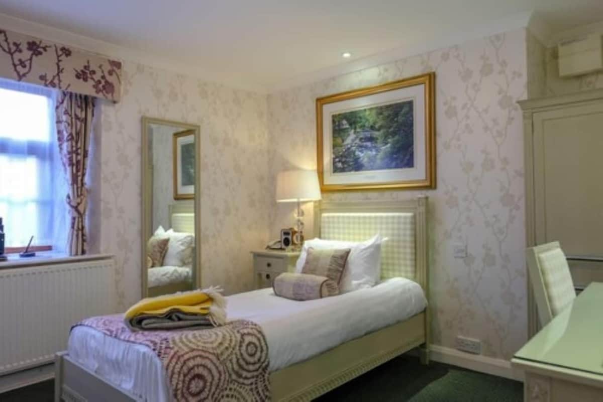 A bedroom with a bed, a bedside table, a lamp, a framed picture on the wall, and a window with curtains.