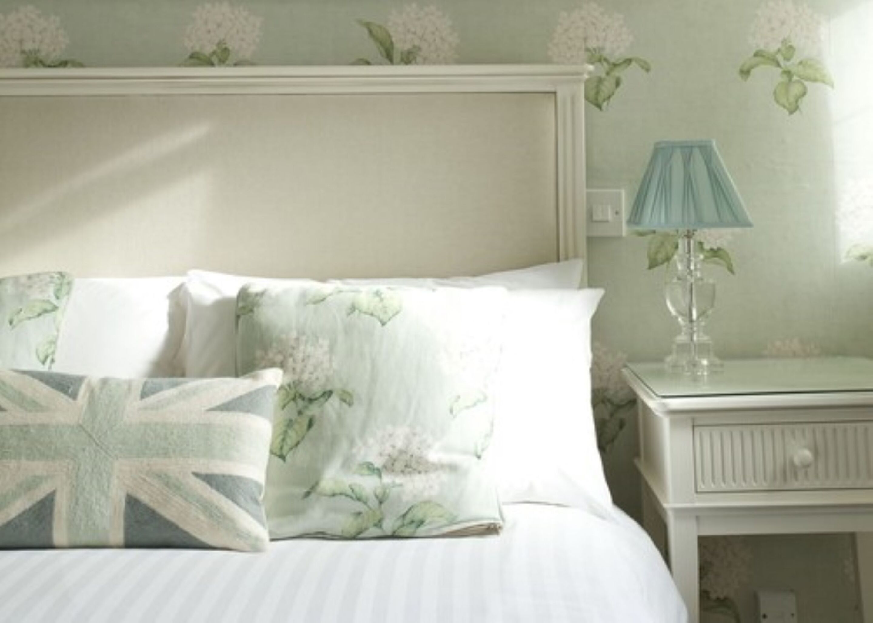 A bed with floral bedding and a Union Jack pillow, a bedside lamp, and a nightstand with a mirror.
