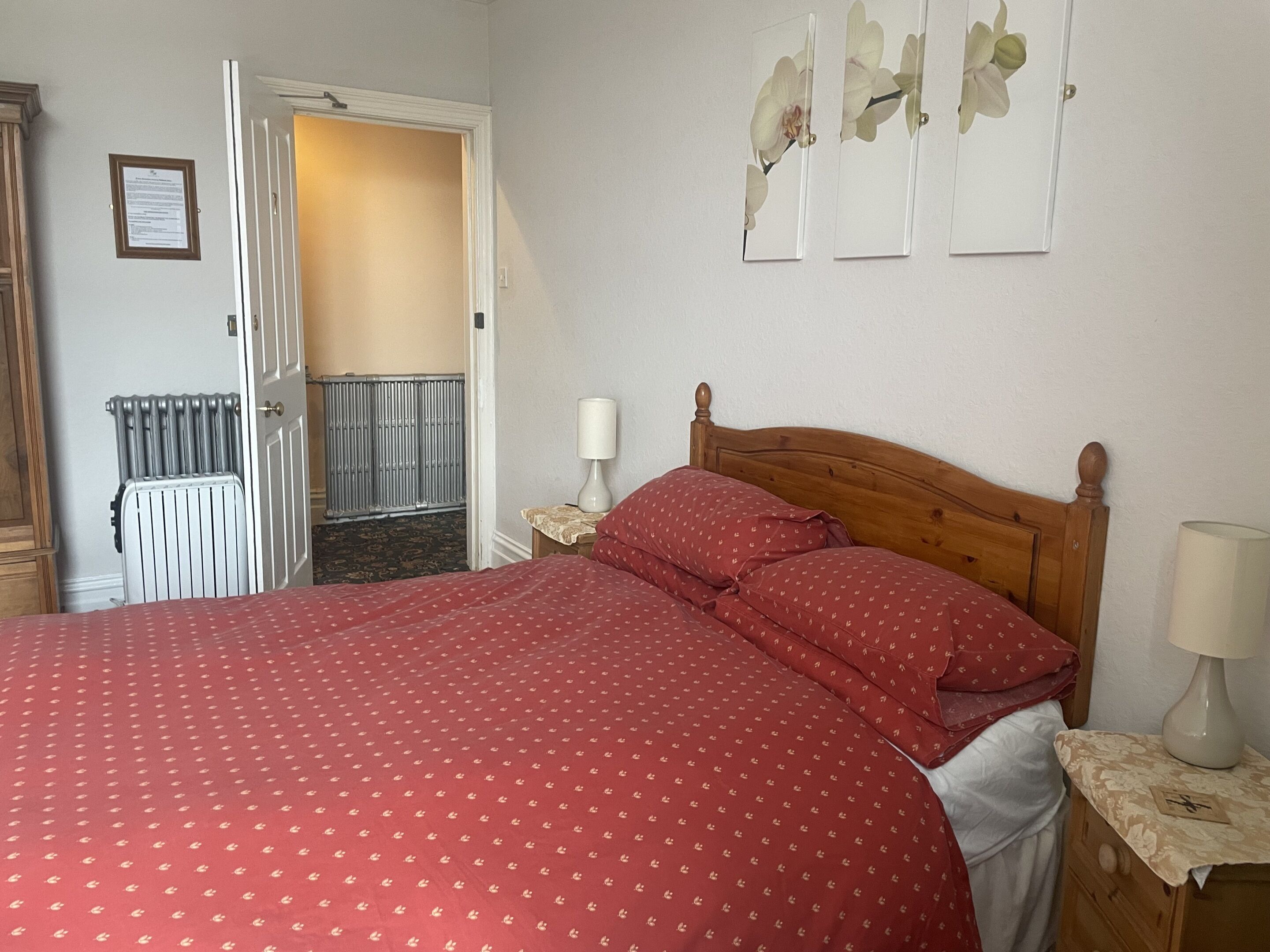 A bedroom with a wooden bed, red bedding, a nightstand with a lamp, a radiator, and a door leading to another room.