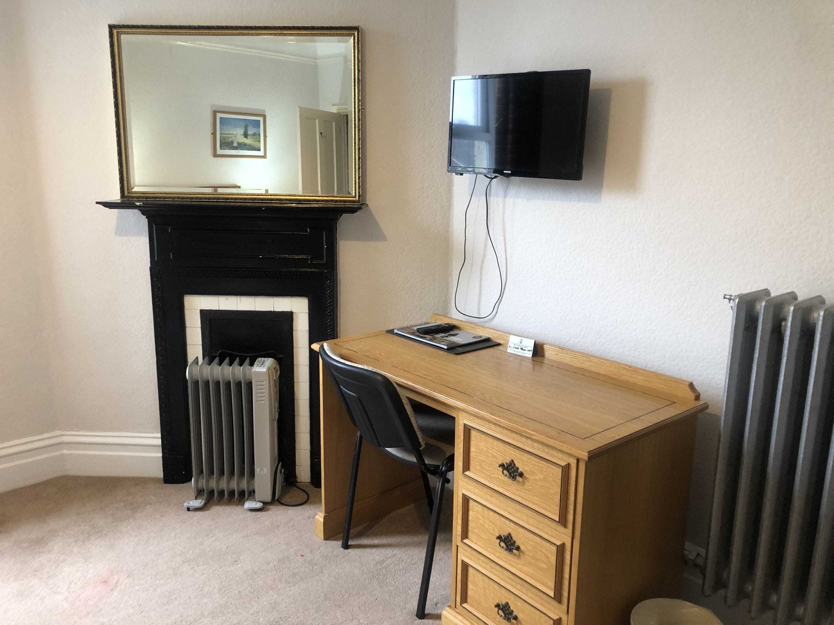 Une pièce avec un bureau en bois, une chaise noire, une cheminée, un radiateur et un téléviseur fixé au mur.