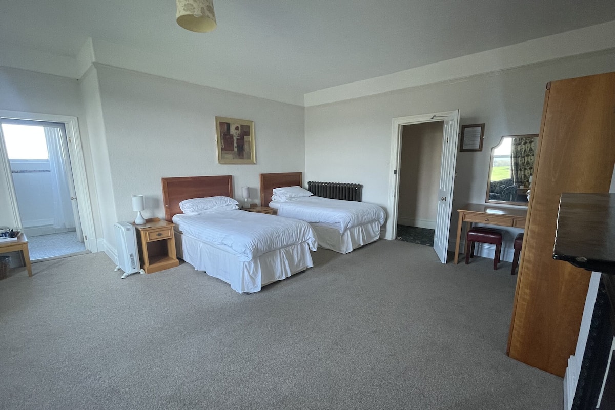 Een hotelkamer met twee bedden, een bureau, een stoel, een radiator en een deur naar een andere kamer.