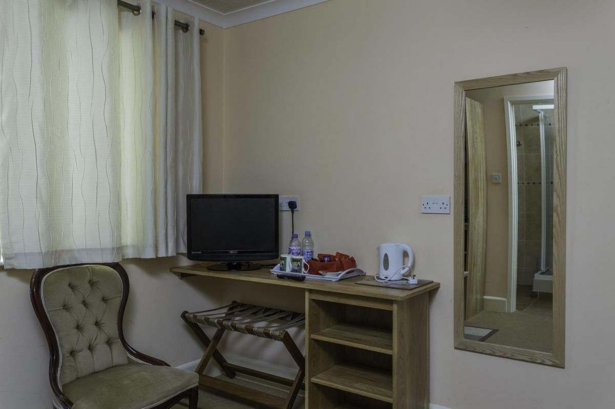 Una habitación con cama, escritorio con computadora, silla, pequeña mesa con hervidor y espejo en la pared.