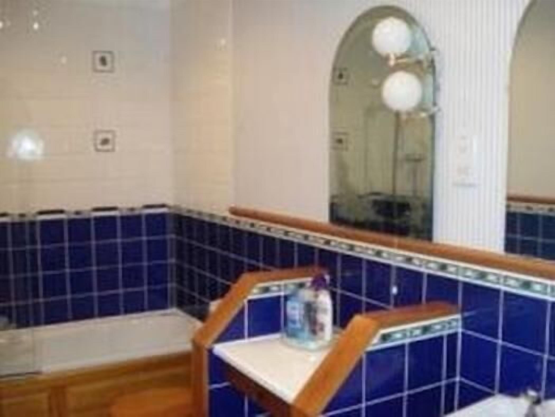 Un baño con azulejos azules, un banco de madera y un espejo con una lámpara redonda.