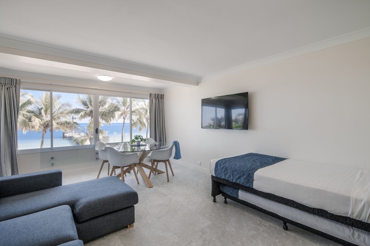 Quarto moderno com cama, televisão, mesa de jantar com cadeiras e vista para palmeiras e mar.