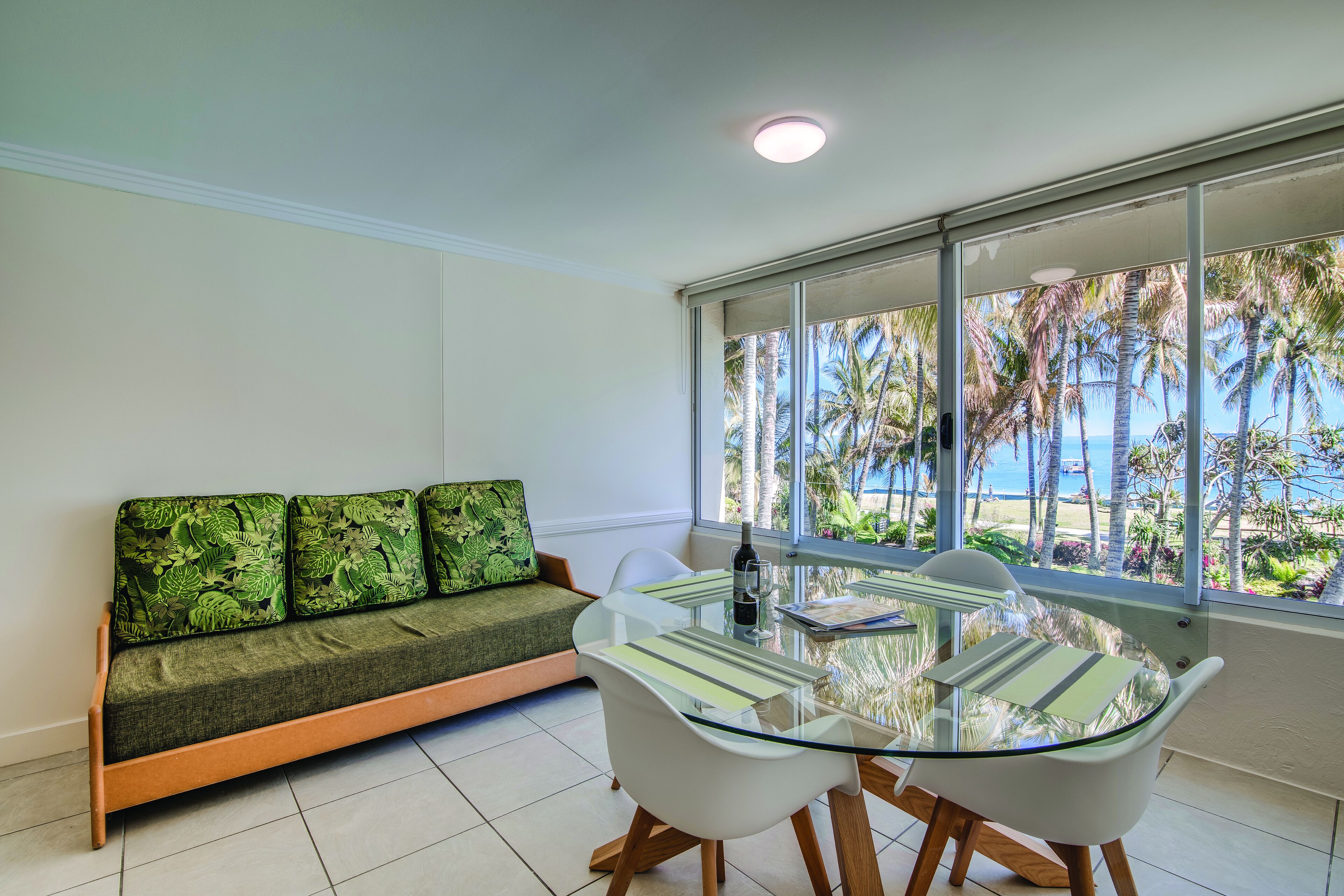 A modern living room with a glass-top dining table, white chairs, and a green sofa with patterned cushions.