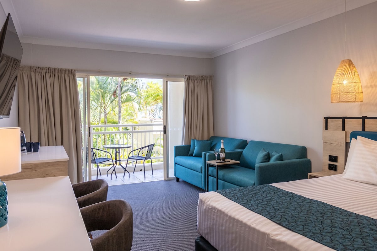 Quarto de hotel com cama, sofá, escrivaninha e varanda com vista para palmeiras.