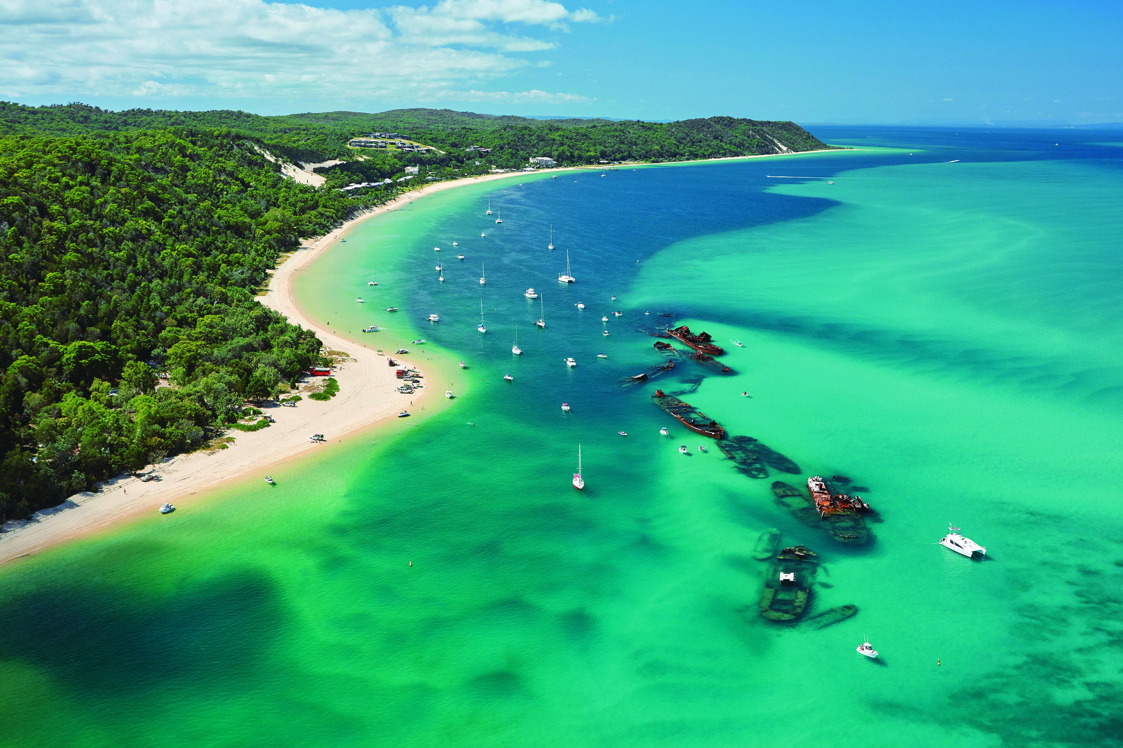 Vista aérea de una zona costera con playa, aguas turquesas cristalinas y varios barcos.