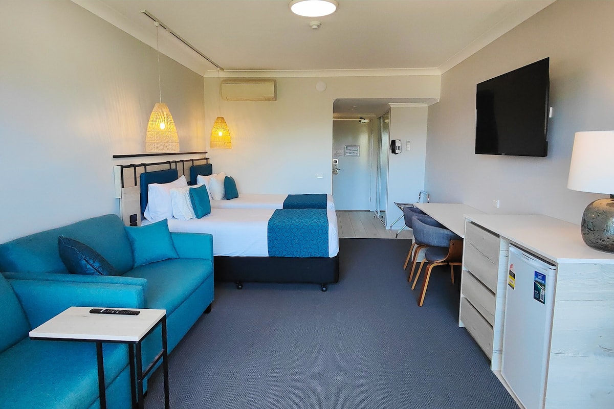 Quarto de hotel com cama, escrivaninha, cadeira, sofá e televisão.