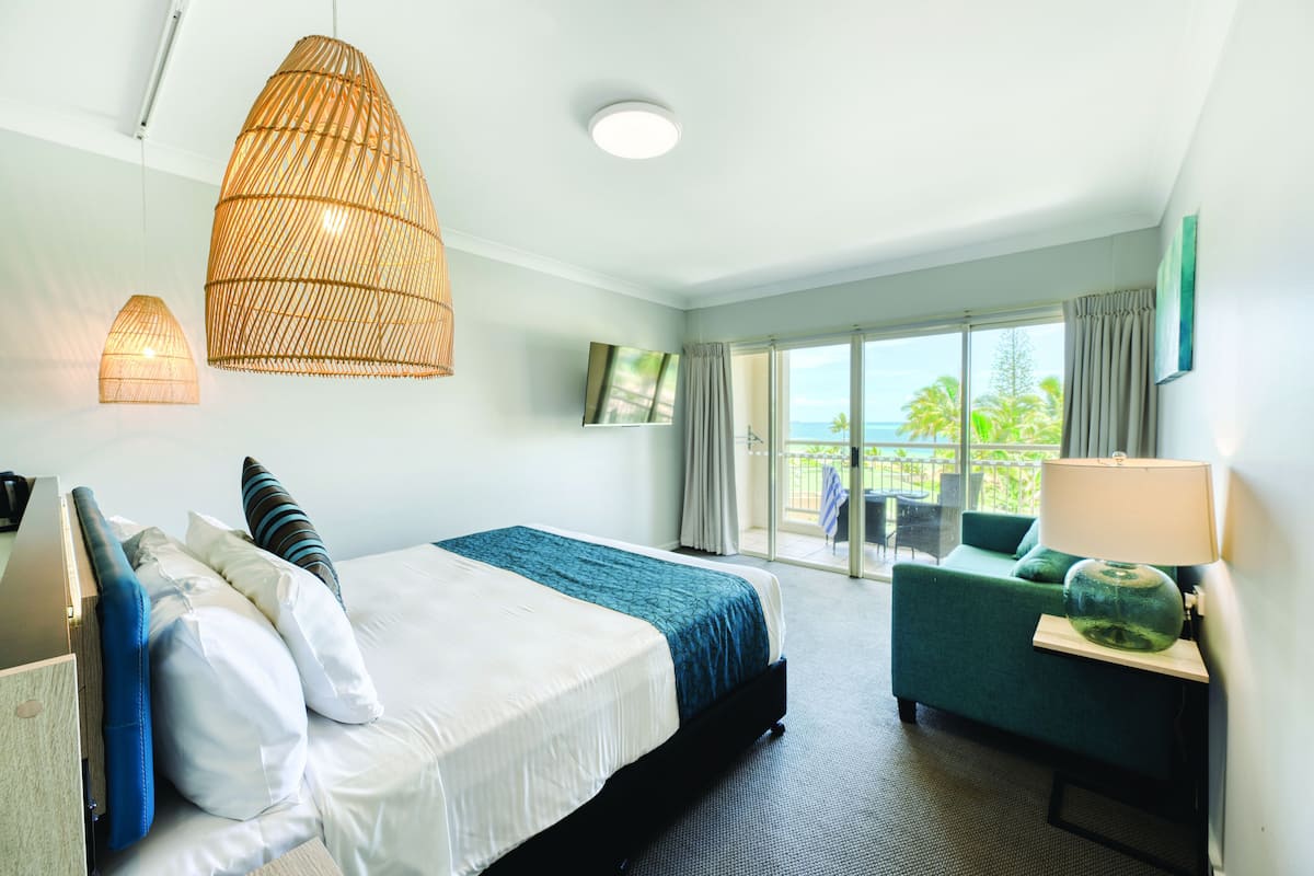 Quarto de hotel com cama, escrivaninha, cadeira, luminária e vista para o exterior.