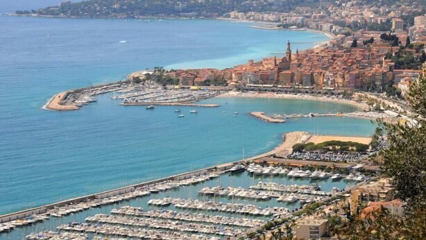 Un paysage urbain côtier avec un port de plaisance rempli de bateaux, un clocher d’église imposant et une zone urbaine densément construite avec des bâtiments aux toits rouges.