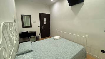 A bedroom with a bed, a desk, a chair, a mirror, a door, and a TV mounted on the wall.