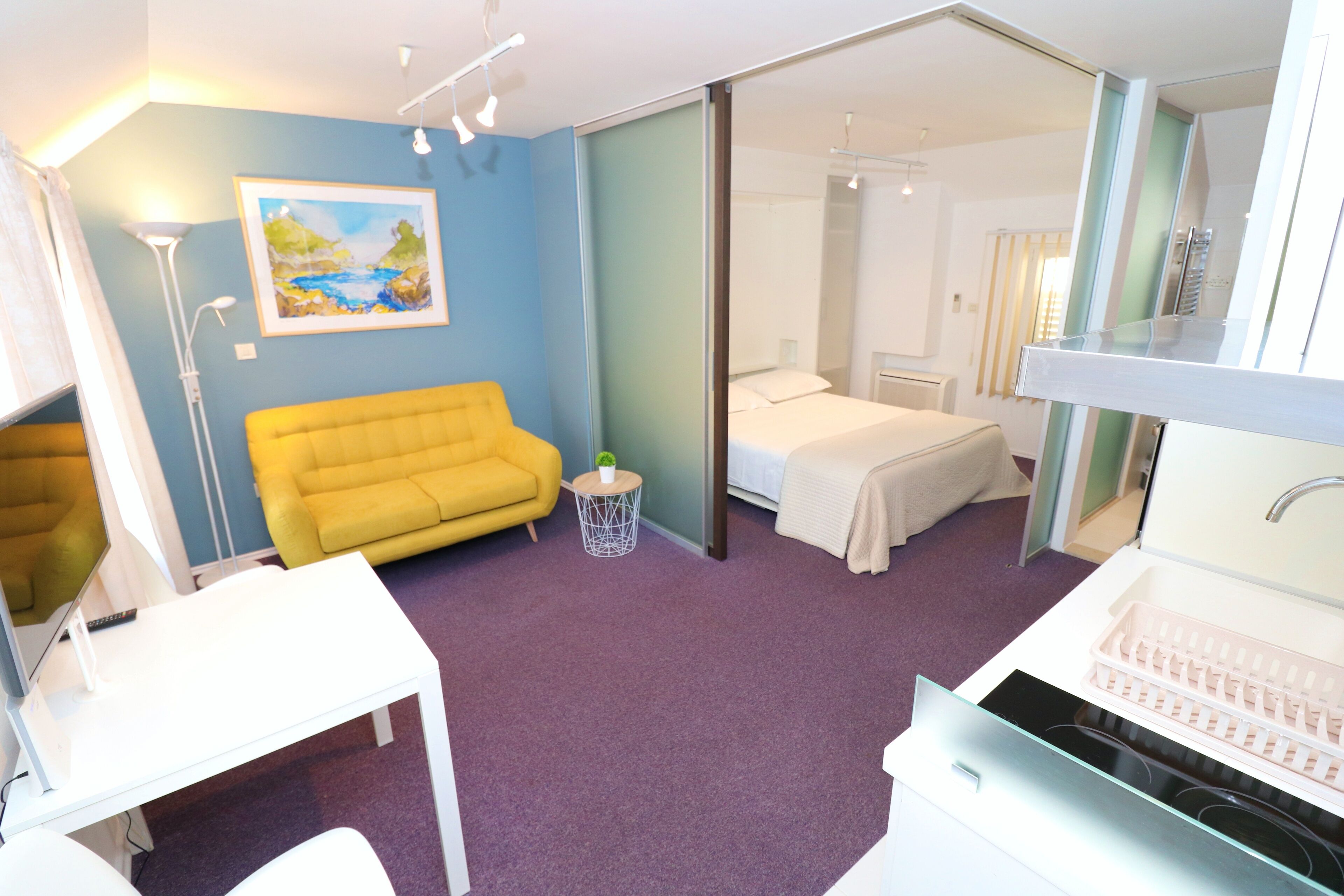 A hotel room with a yellow sofa, a bed, a white desk, and a bathroom with a sink and mirror.