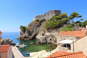A coastal town with a historic stone castle on a cliff, clear blue sea, and traditional buildings with red-tiled roofs.