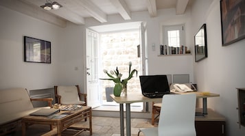 A room with a wooden floor, a white sofa, a small table with a laptop, a chair, a window with books on a shelf, and a door leading outside.
